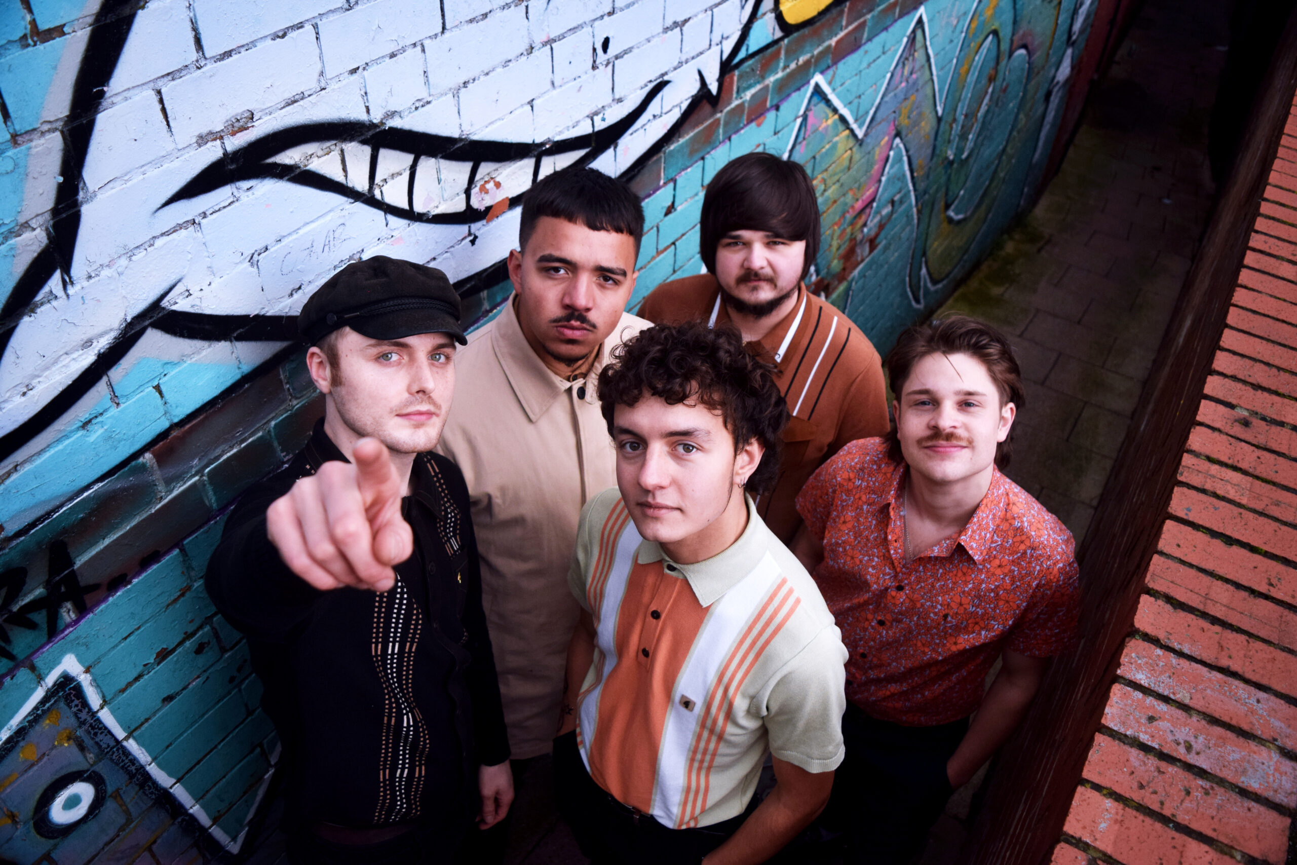 5 people looking up at the camera. The person on the far left is pointing into the lens of the camera. There is a wall behind them covered in graffiti art.