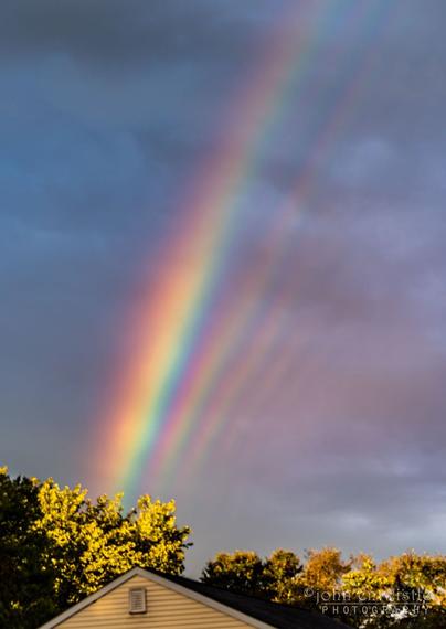 The top of a house is shown beneath a dark and cloudy sky. Many rainbow stripes are seen above the house.