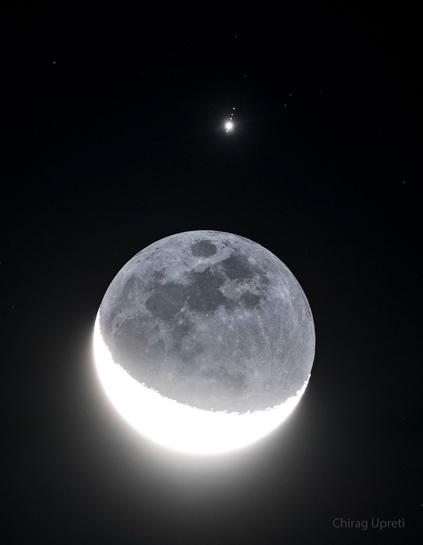In front of a dark background an image of Earth's Moon appears with a lower crescent brightly lit. The rest of the Moon is also faintly visible. Above the Moon is a bright point of  light surrounded by four fainter points of light in row.