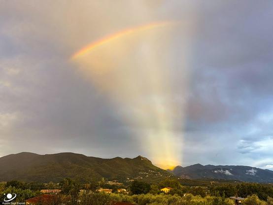 Hills are seen in the distance below a partly cloudy sky. A group of orange rays appears to eminate from one area behind the hills. Across this fan of rays, near the top of the image, a short rainbow can be seen.