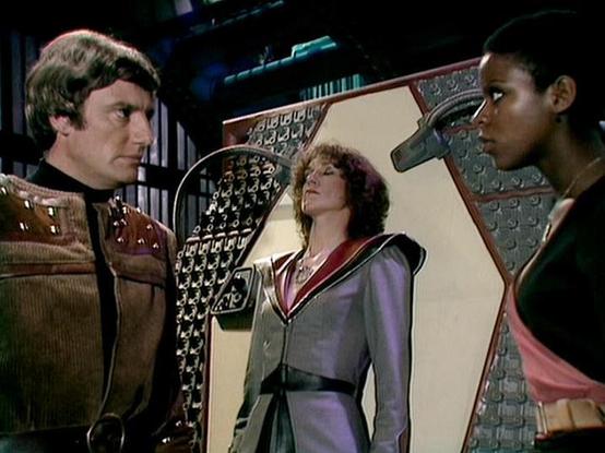 Claude Sonnet 5.5 describes the image as: "This still shows a tense moment on a low-budget, late-1970s/early-1980s British sci-fi set, with the soft, slightly grainy look of studio video. Taking your word that the cast includes Paul Darrow, Jan Chappell and Josette Simon, I won't match names to individual faces. On the left, a man with tousled brown hair stands in a brown corduroy-style jacket with metallic studs on the shoulders, a black high-collared undershirt and a large belt buckle. His expression is stern as he looks toward the others. In the center, a woman with curly auburn hair stands rigidly upright against an angular panel with her eyes closed. She wears a tailored silver-gray jacket with red trim, sharply structured shoulders and a pendant necklace, and she looks entranced or incapacitated. On the right, a woman with closely cropped dark hair is seen in profile, gazing intently. She wears a dark top with a salmon-pink panel and a delicate necklace. Behind them is a cream geometric panel framed by patterned metallic lattice, set against a dark, industrial-looking backdrop. Together these give the scene a futuristic, suspenseful at"