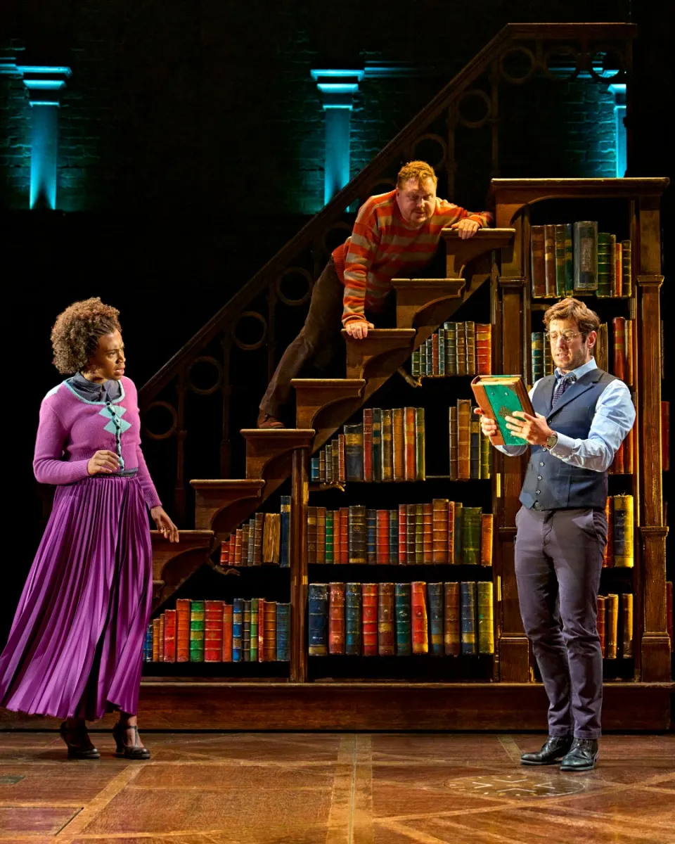 Three actors on stage with a bookshelf backdrop.