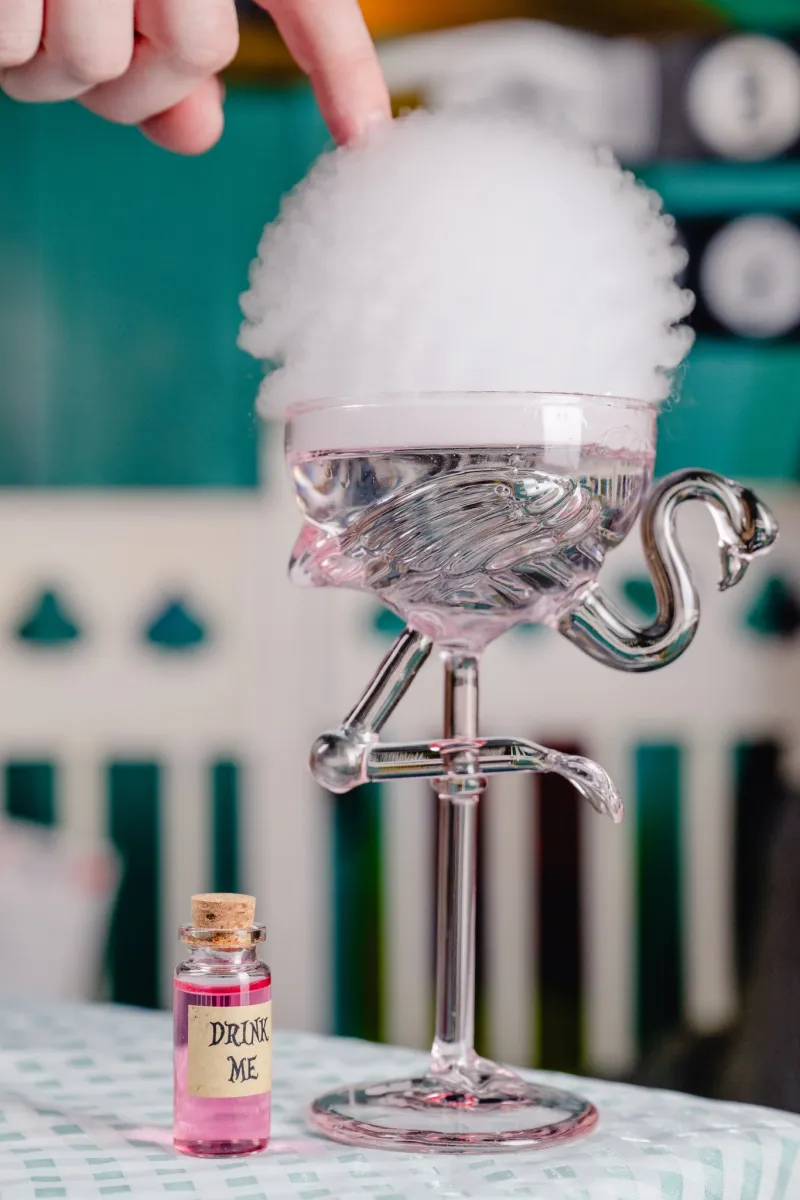 Glass with flamingo stem and smoke, small bottle labeled 'Drink Me'.
