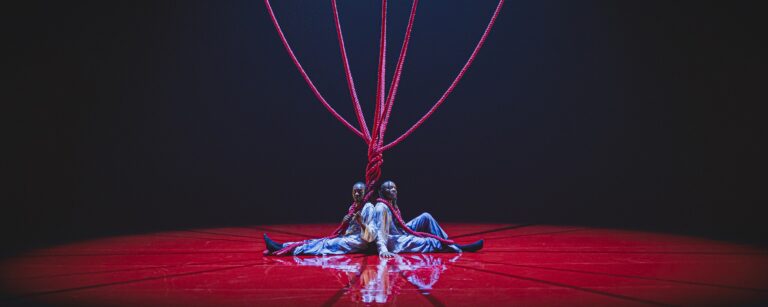 Two black men sitting on a red floor with red ropes twisted around them and rising upwards above them