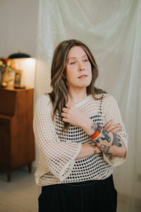 A woman with long brown hair looks calmly off camera. She wears a loose knit pale jumper and a number of tattoos are visible on her arm.